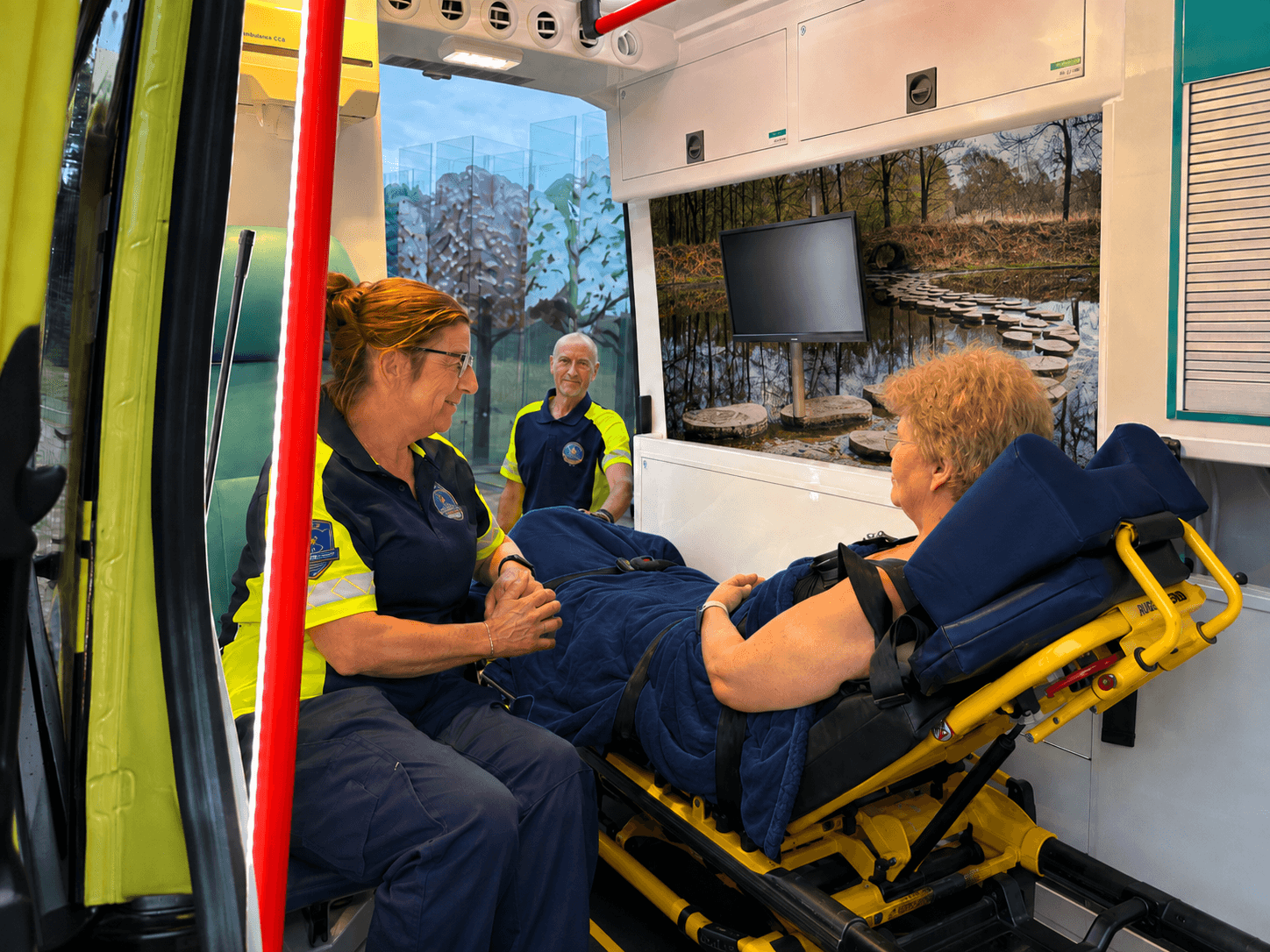 Twee vrijwilligers van de WensAmbulance in gele hesjes praten met een gast op de brancard in de ambulance