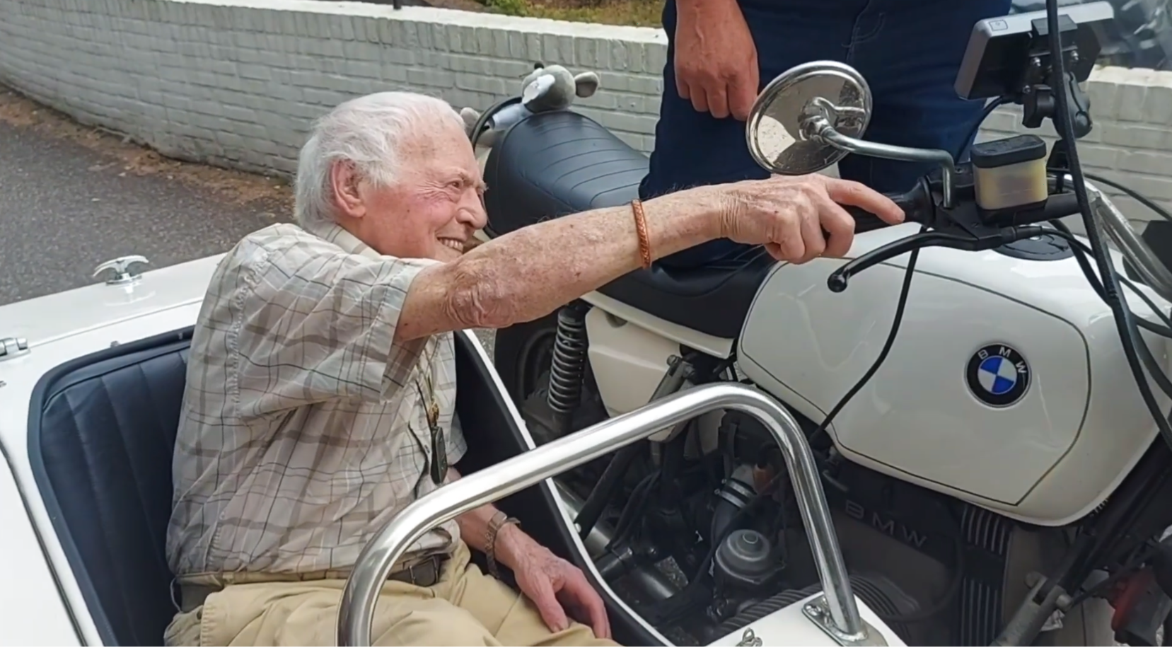 Een glimlachende oudere man in een wit zijspan reikt naar het stuur van een witte BMW-motor