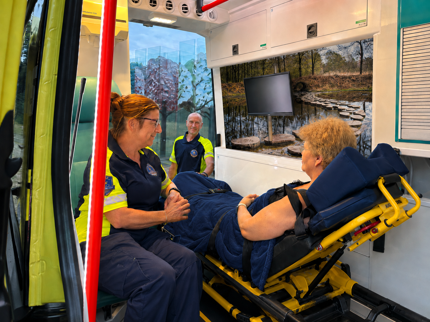 Twee vrijwilligers van de WensAmbulance in gele hesjes praten met een gast op de brancard in de ambulance