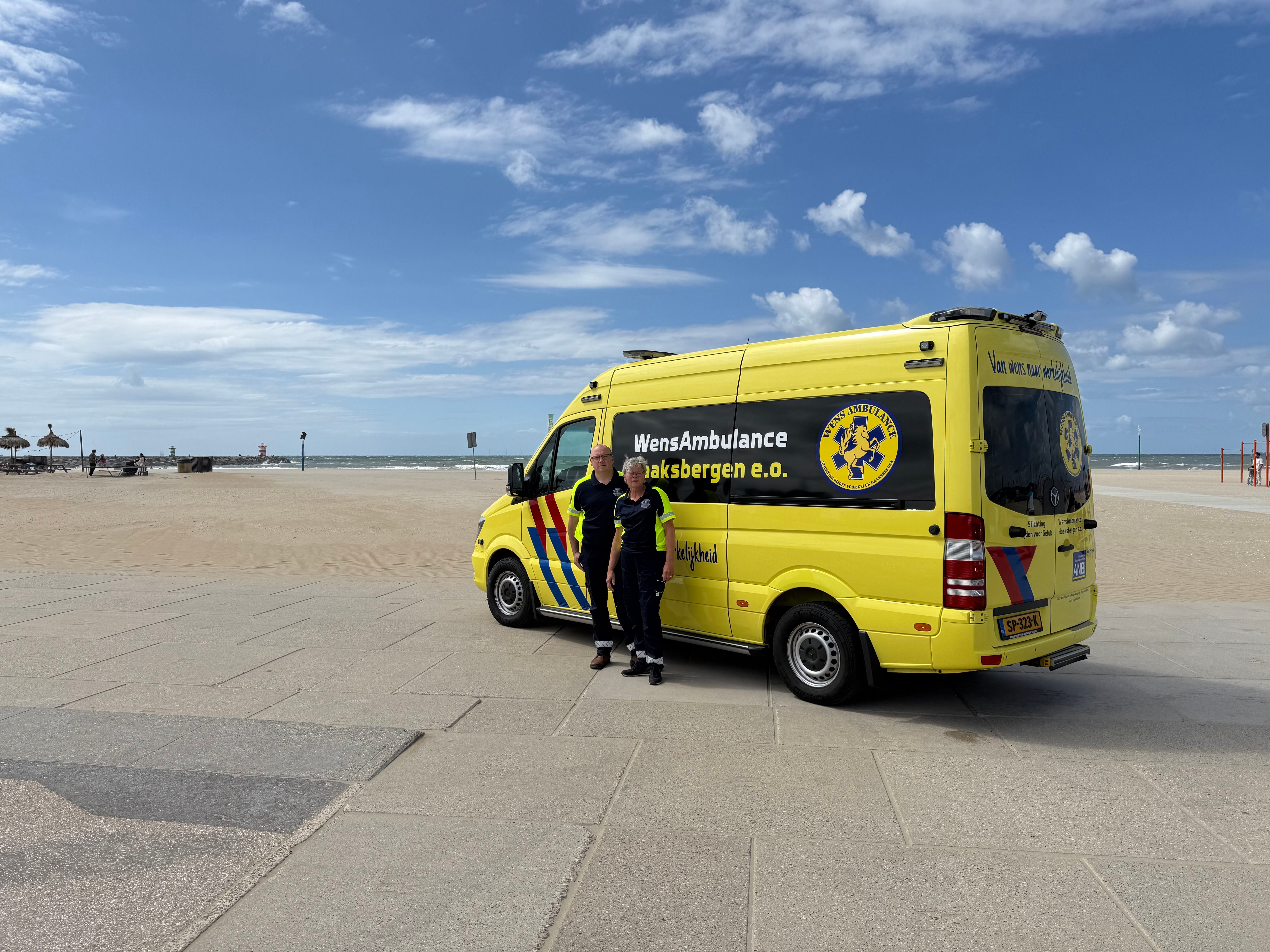 De gele WensAmbulance van Stichting Rijden voor Geluk op de strandboulevard met twee vrijwilligers ernaast